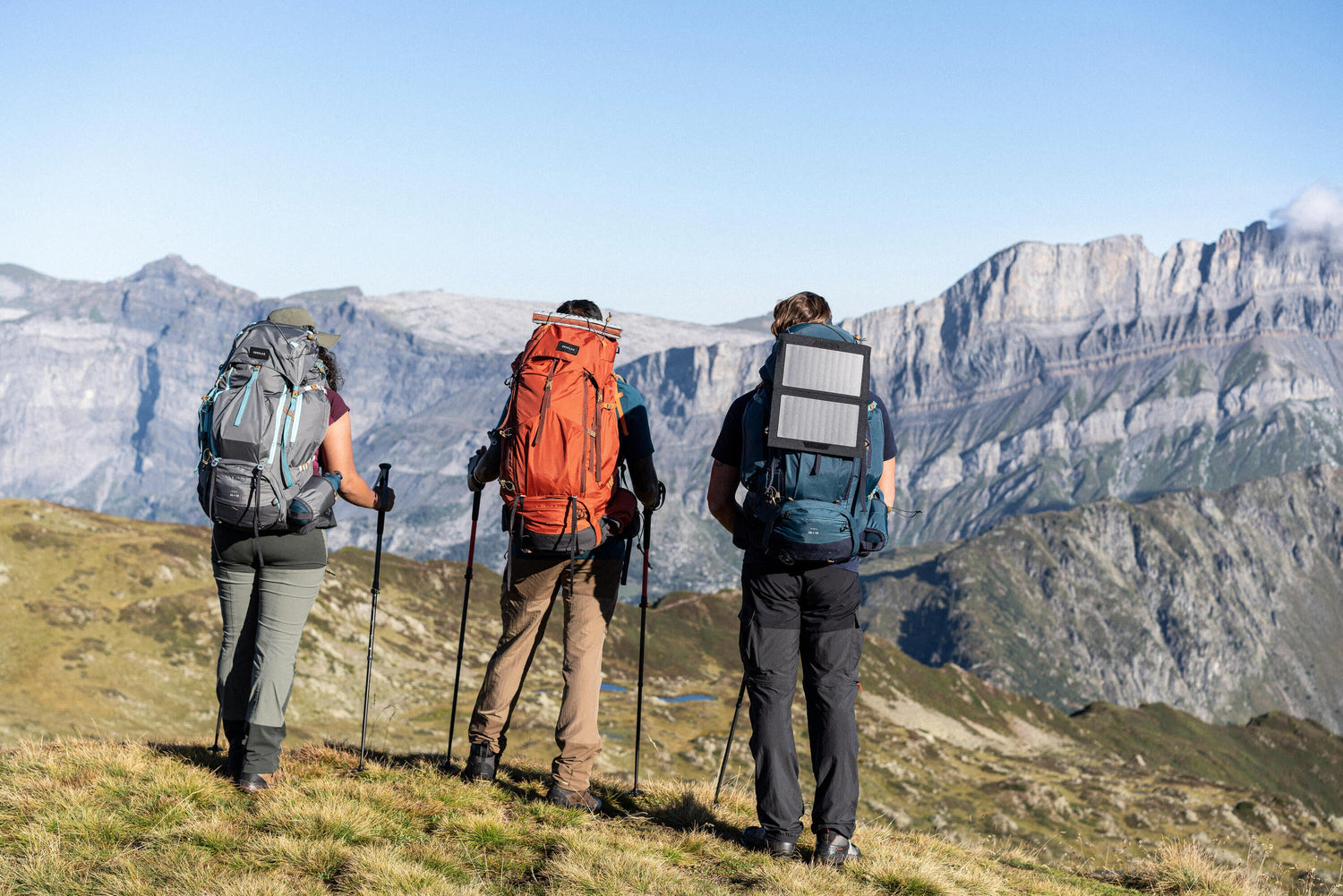 Three hikers with large backpacks trekking through a mountain trail with trekking poles, overlooking a scenic alpine landscape.