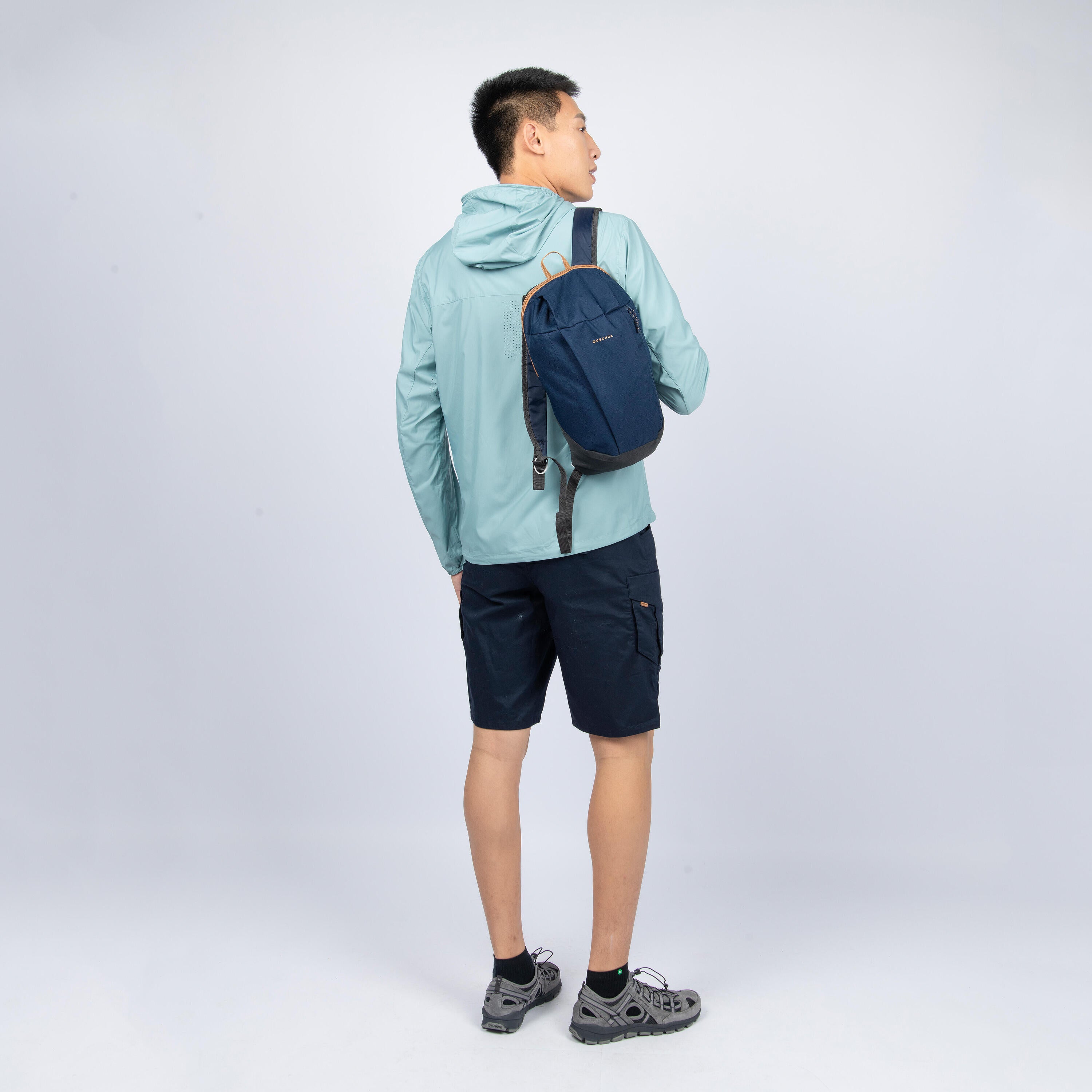 Person wearing a light blue jacket and dark shorts with a navy backpack on a white background