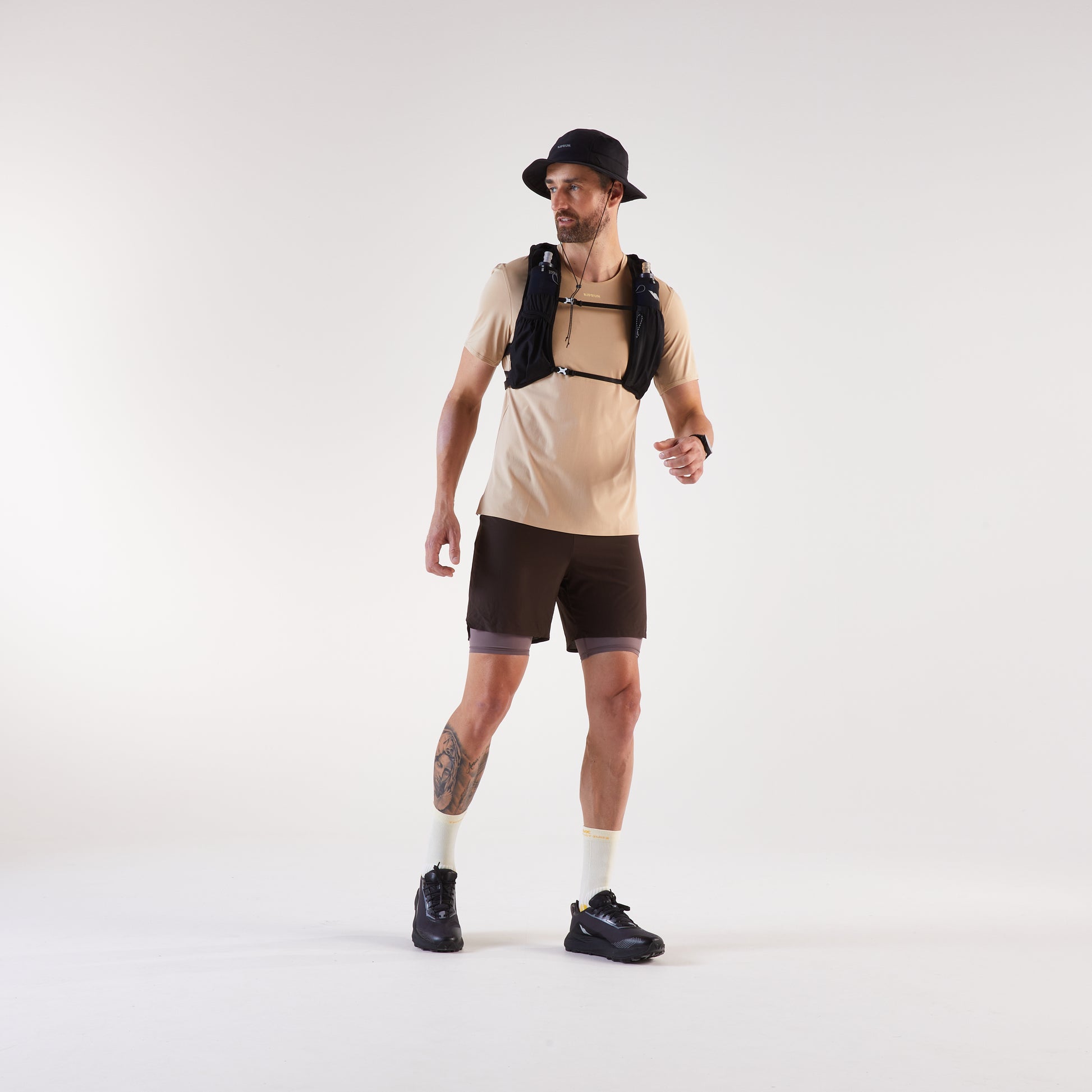 Man wearing a beige t-shirt, black shorts, and a black backpack on a white background