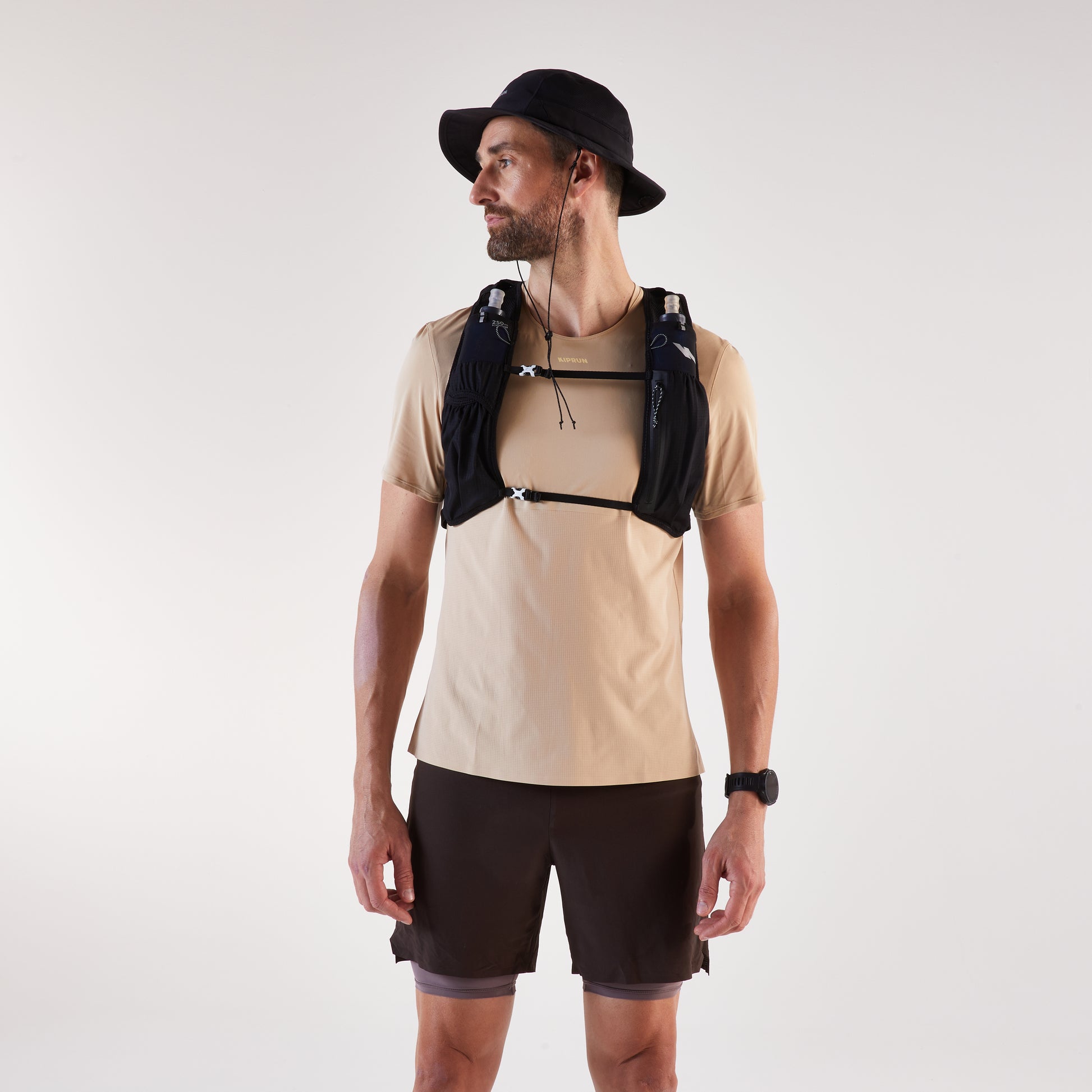 Man wearing a beige t-shirt, black shorts, and a black hat with a hydration vest on a white background