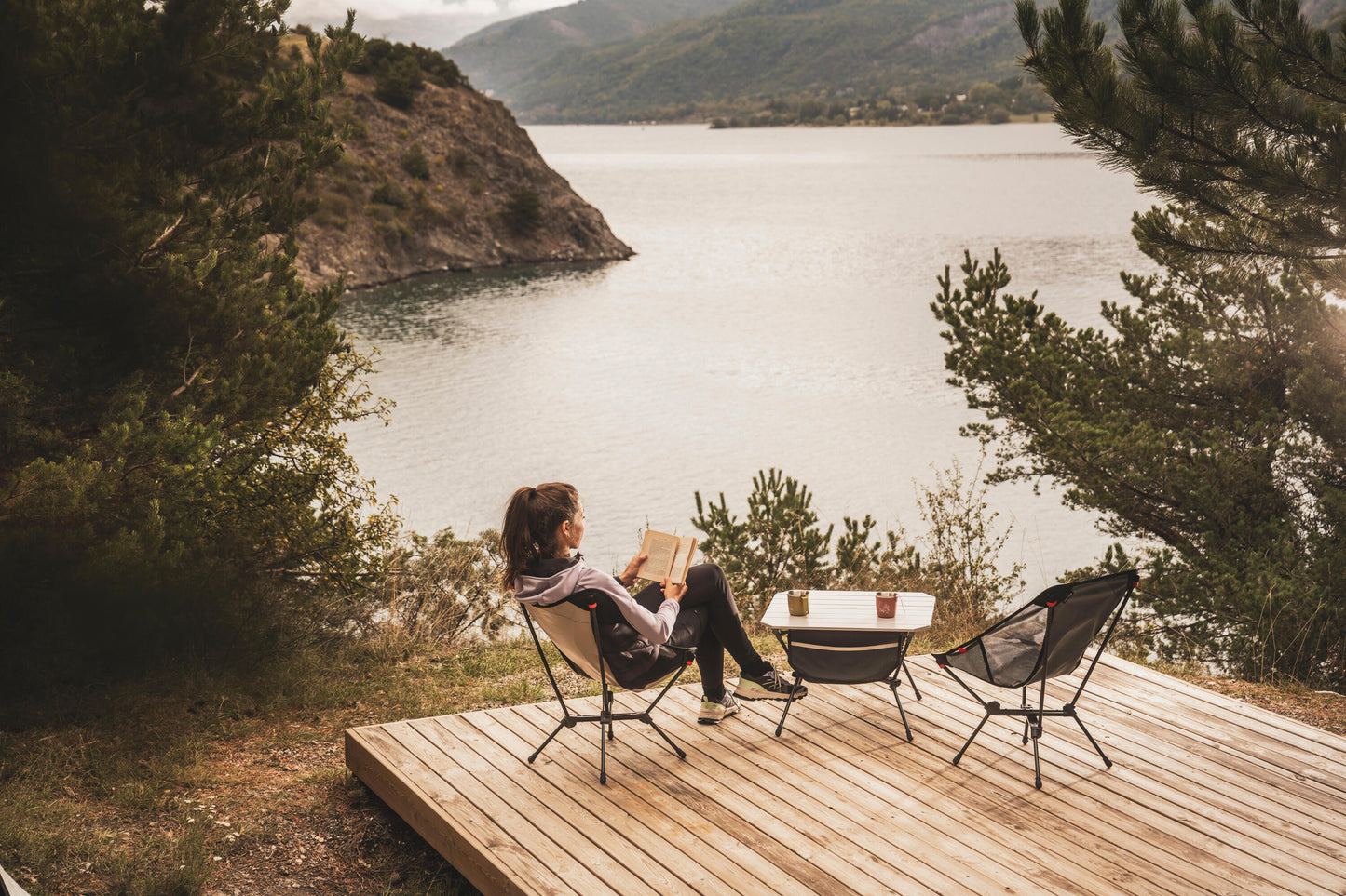 Person reading a book on a wooden deck overlooking a lake with mountains in the background