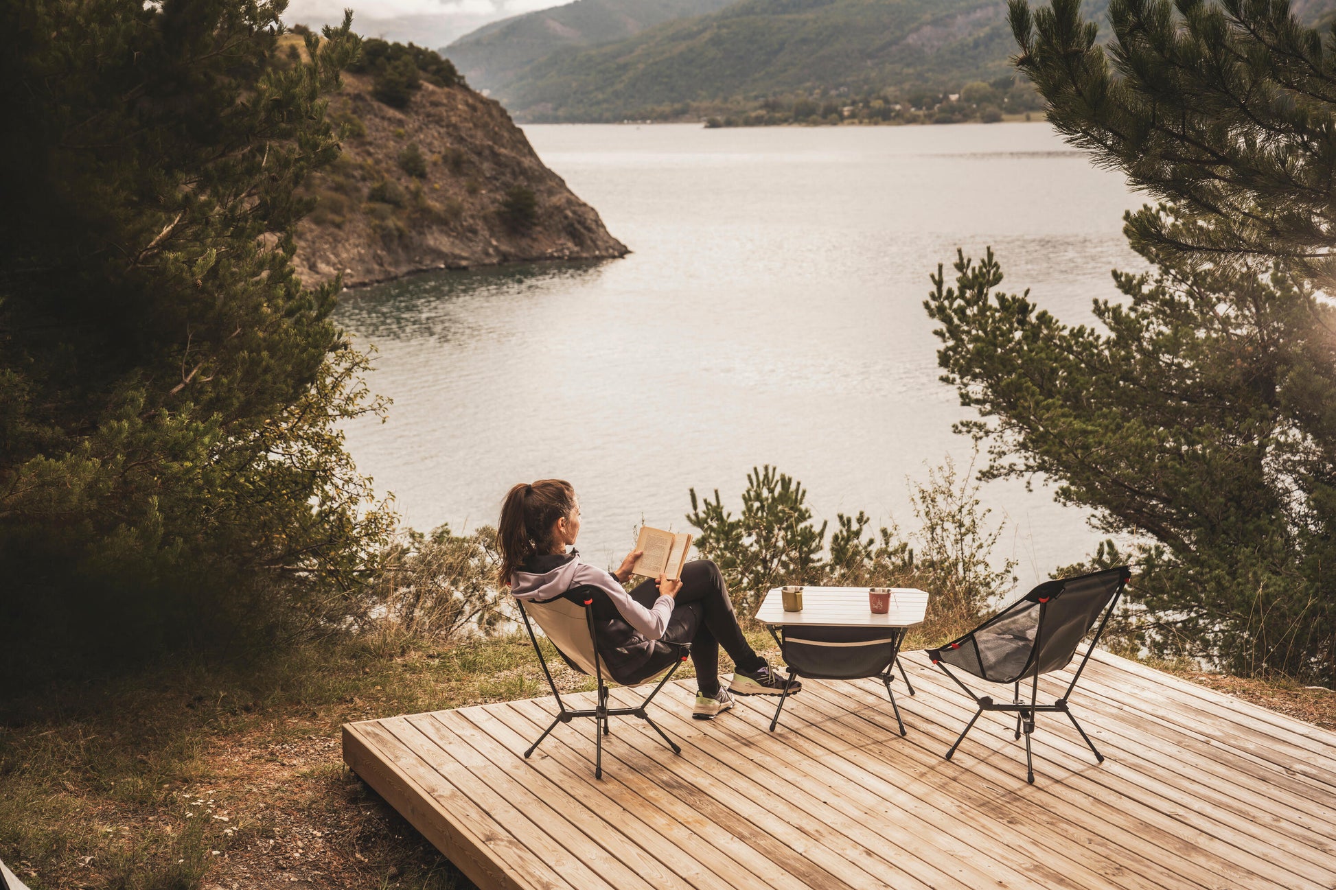 Person reading a book on a wooden deck overlooking a lake with mountains in the background
